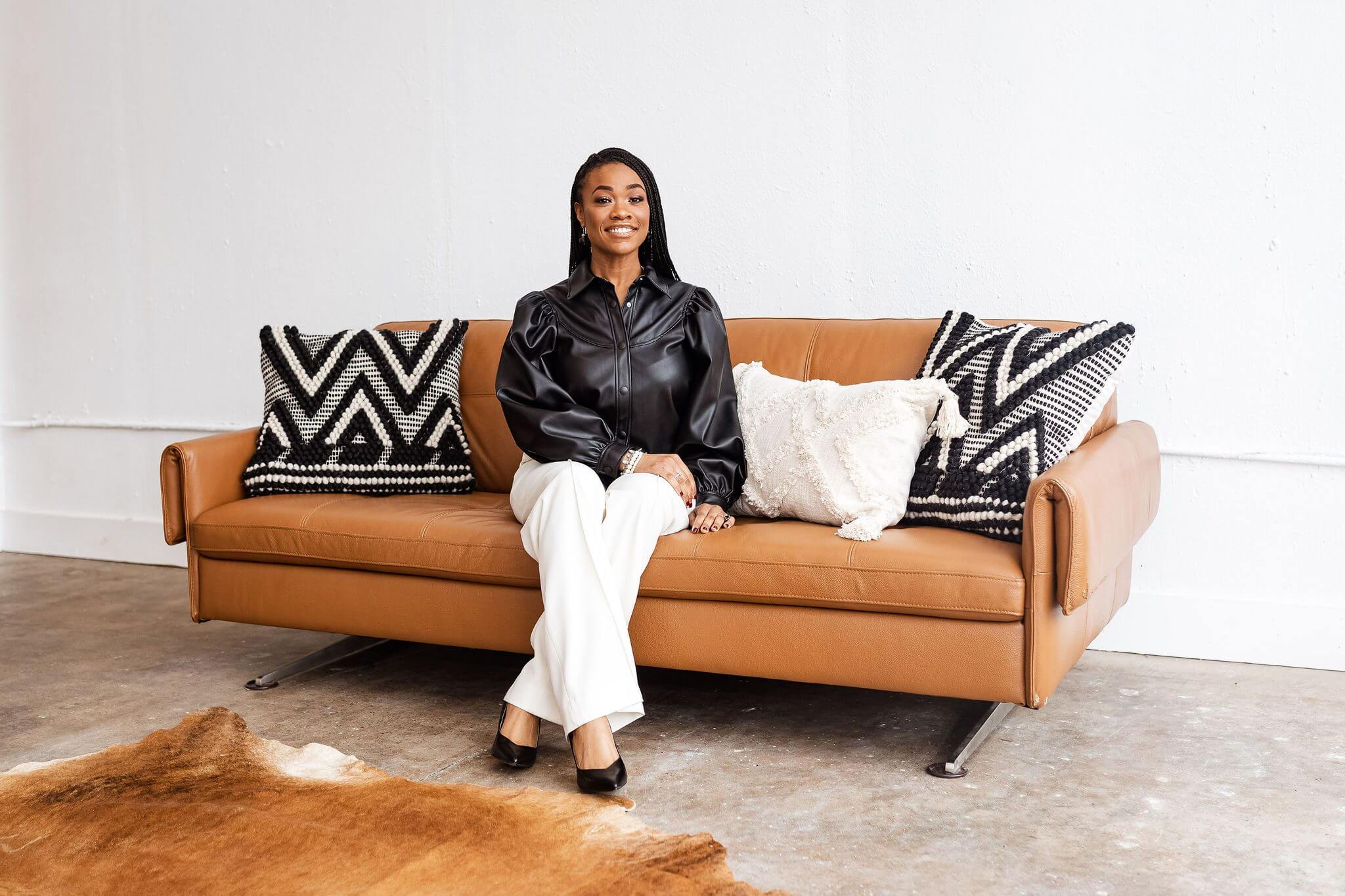 A woman in a black blouse and white pants sitting on a brown sofa.